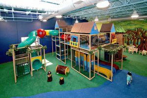 Two level indoor playground with multiple slides and a forest background with durable indoor playground carpeting that has cushioned floors.