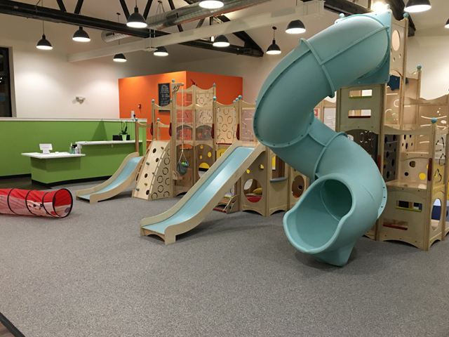 Compliant playground carpeting that meets ADA requirements and made to withstand heavy foot traffic with a large wooden play structure and three blue slides.