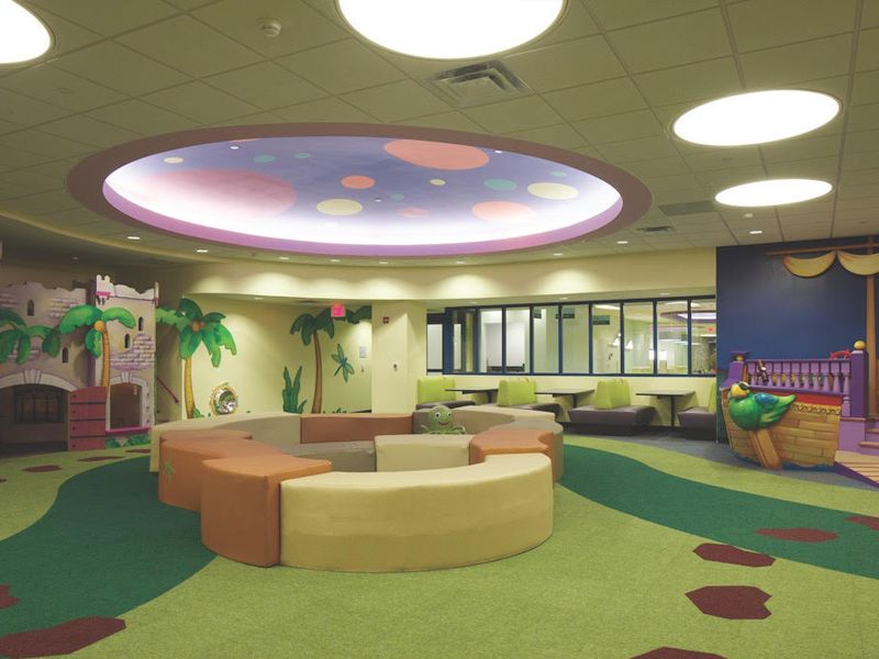 Compliant playground padding with a custom printed green carpeting with brown stepping stones inside a hospital indoor play center with a castle and ship play structure.