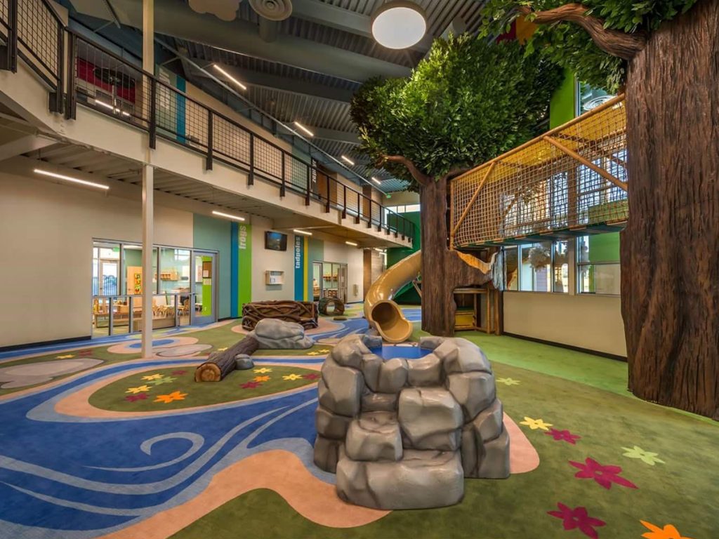 Carpet with custom print for an indoor religious play center that is compliant with playground fall height requirements. There are several play structures including a log, and a slide from the trees.